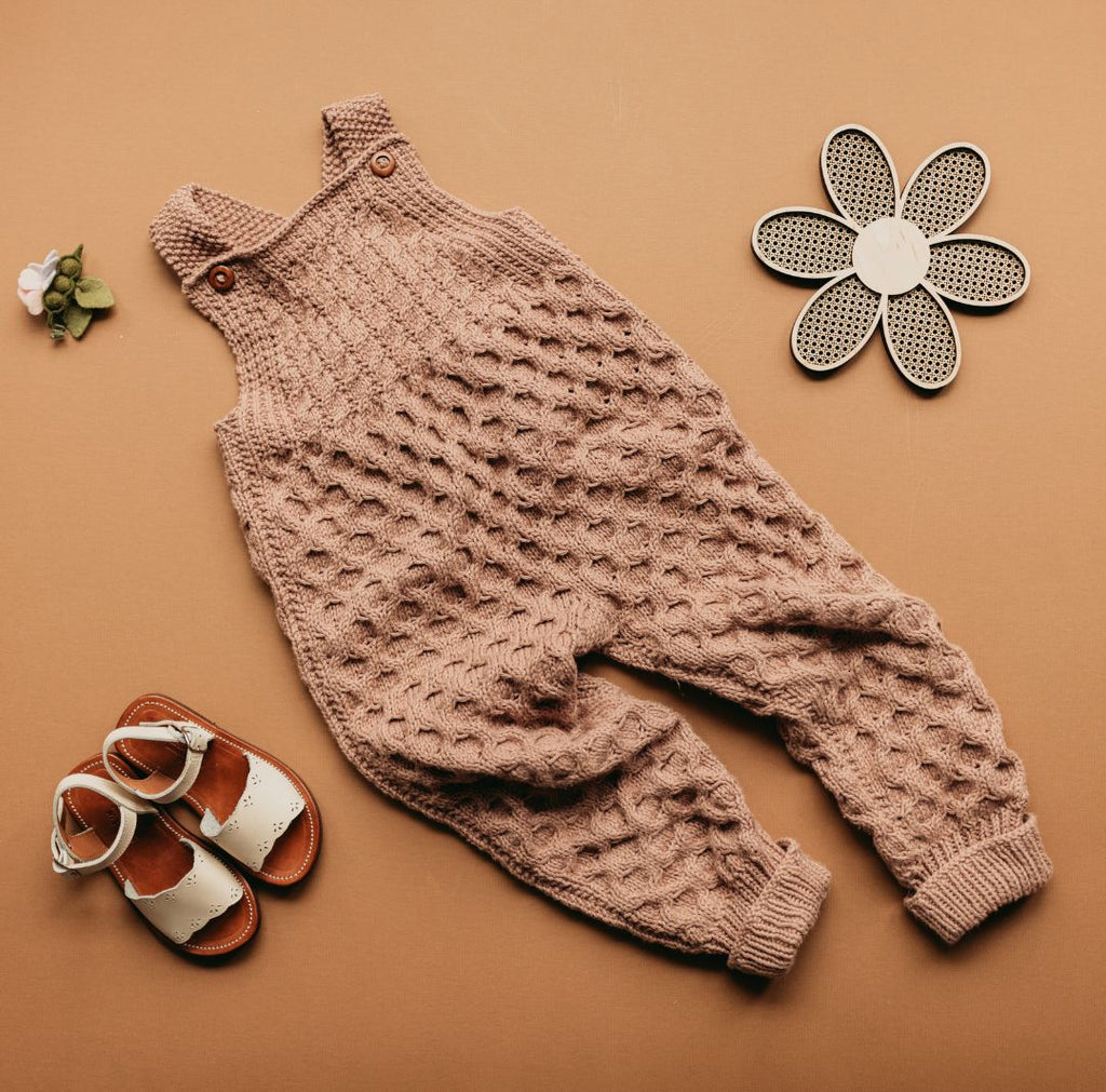 Hand-knit honeycomb romper in light brown, styled with white leather sandals and a wooden flower on a warm tan background