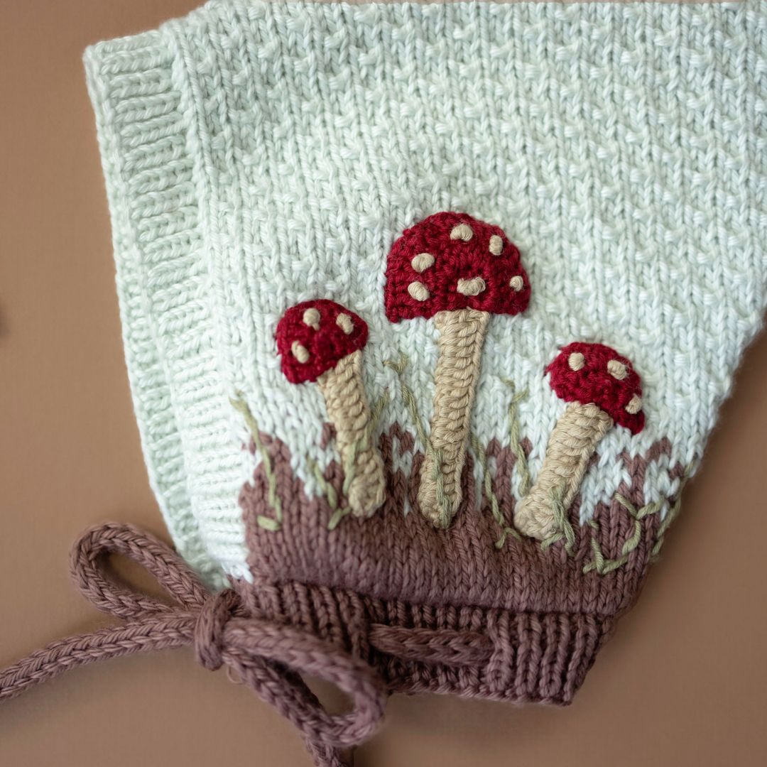 Close-up flat lay of hand-knit mushroom pixie hat in creamy green cotton bamboo showing hand-crocheted mushroom motif detail.