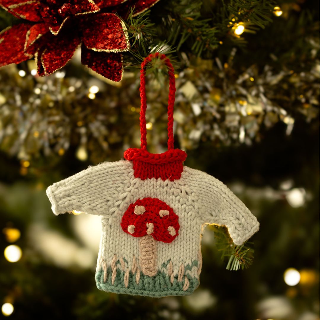 Close-up of hand-knit mushroom sweater ornament in creamy light green cotton showing hand-crocheted mushroom motif detail, eco-friendly holiday gift.