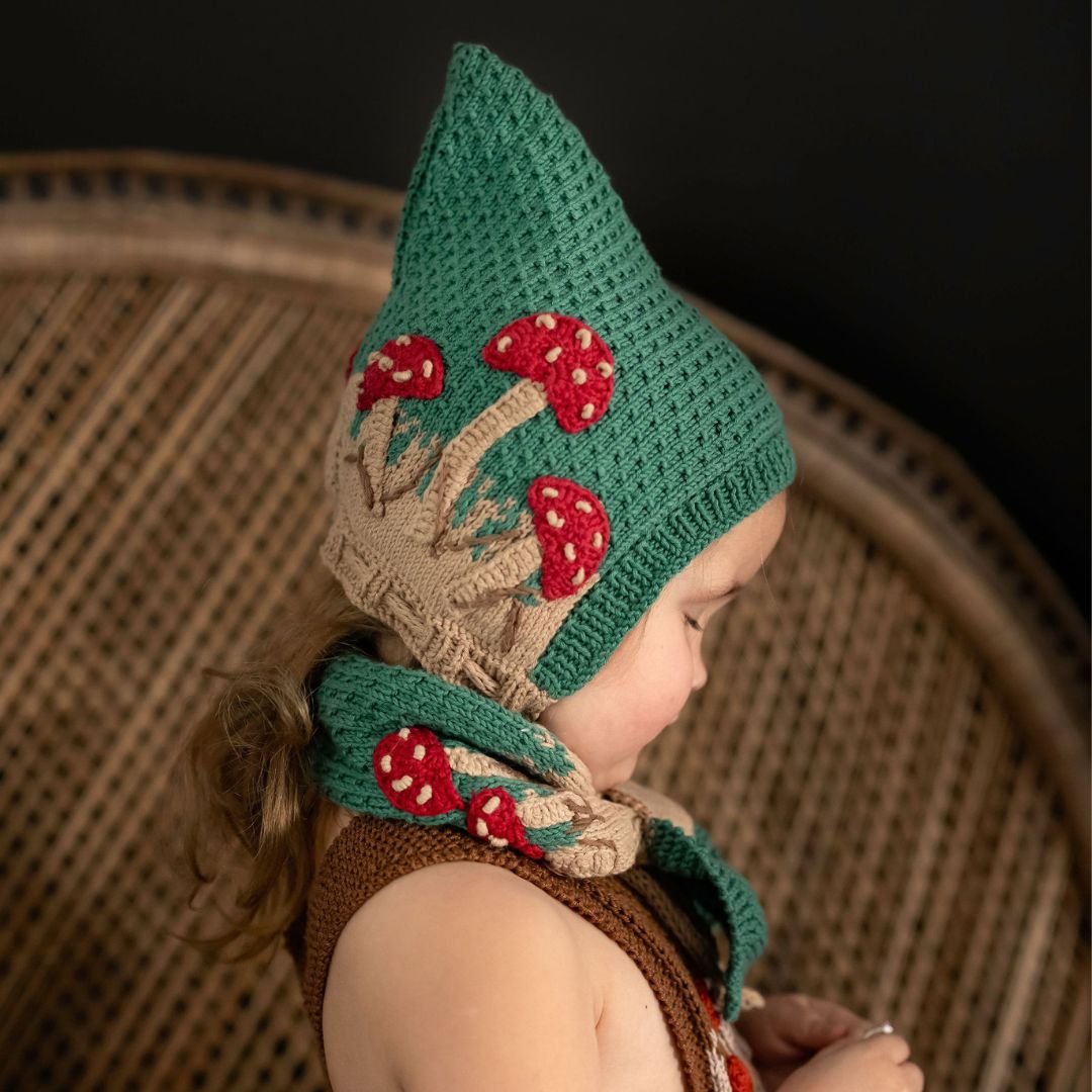 Toddler wearing hand-knit mushroom pixie hat in hunter green cotton bamboo, gender-neutral baby pixie hat with hand-crocheted mushroom motifs.