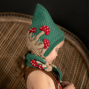 Toddler wearing hand-knit mushroom pixie hat in hunter green cotton bamboo, gender-neutral baby pixie hat with hand-crocheted mushroom motifs.
