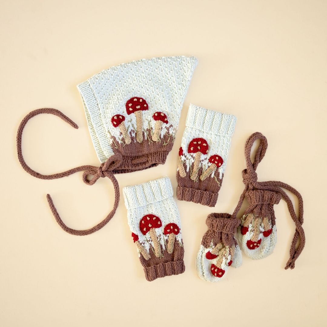 Flat lay of hand-knit accessories set in creamy light green cotton bamboo, featuring pixie hat, leg warmers and mittens with hand-crocheted mushroom motifs.
