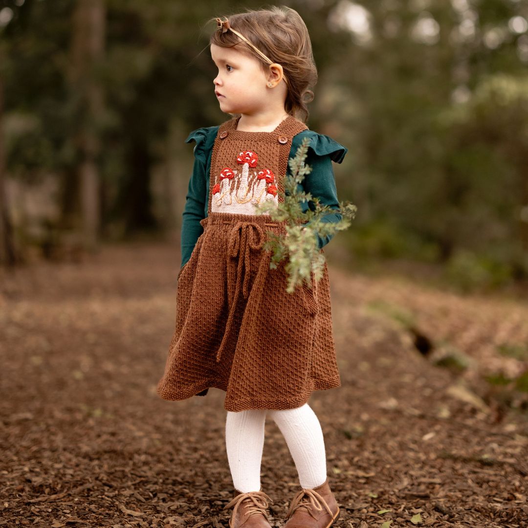 Toddler girl wearing hand-knit mushroom pinafore in chocolate brown cotton bamboo with hidden pockets and adjustable straps.