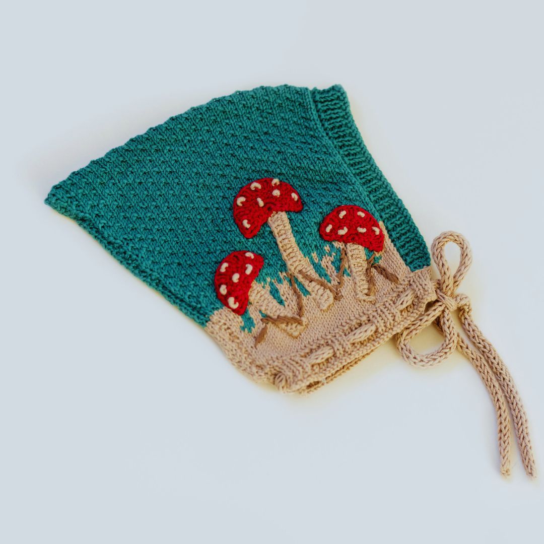 Flat lay of hand-knit mushroom pixie hat in hunter green cotton bamboo with hand-crocheted mushroom motifs, heirloom quality baby pixie hat.