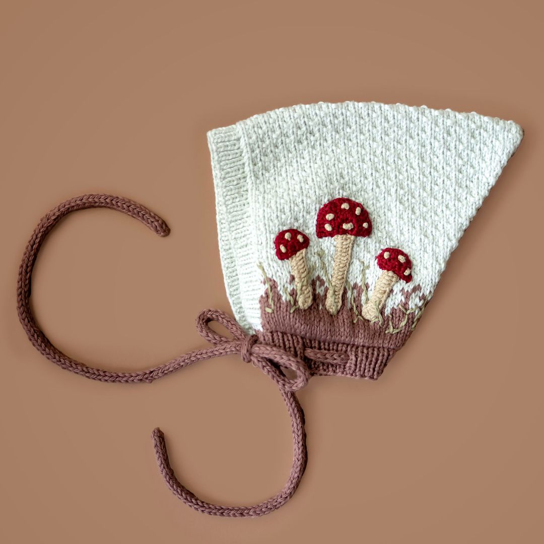 Flat lay of hand-knit mushroom pixie hat in creamy light green cotton bamboo with hand-crocheted mushroom motifs, artisan-made baby pixie hat.