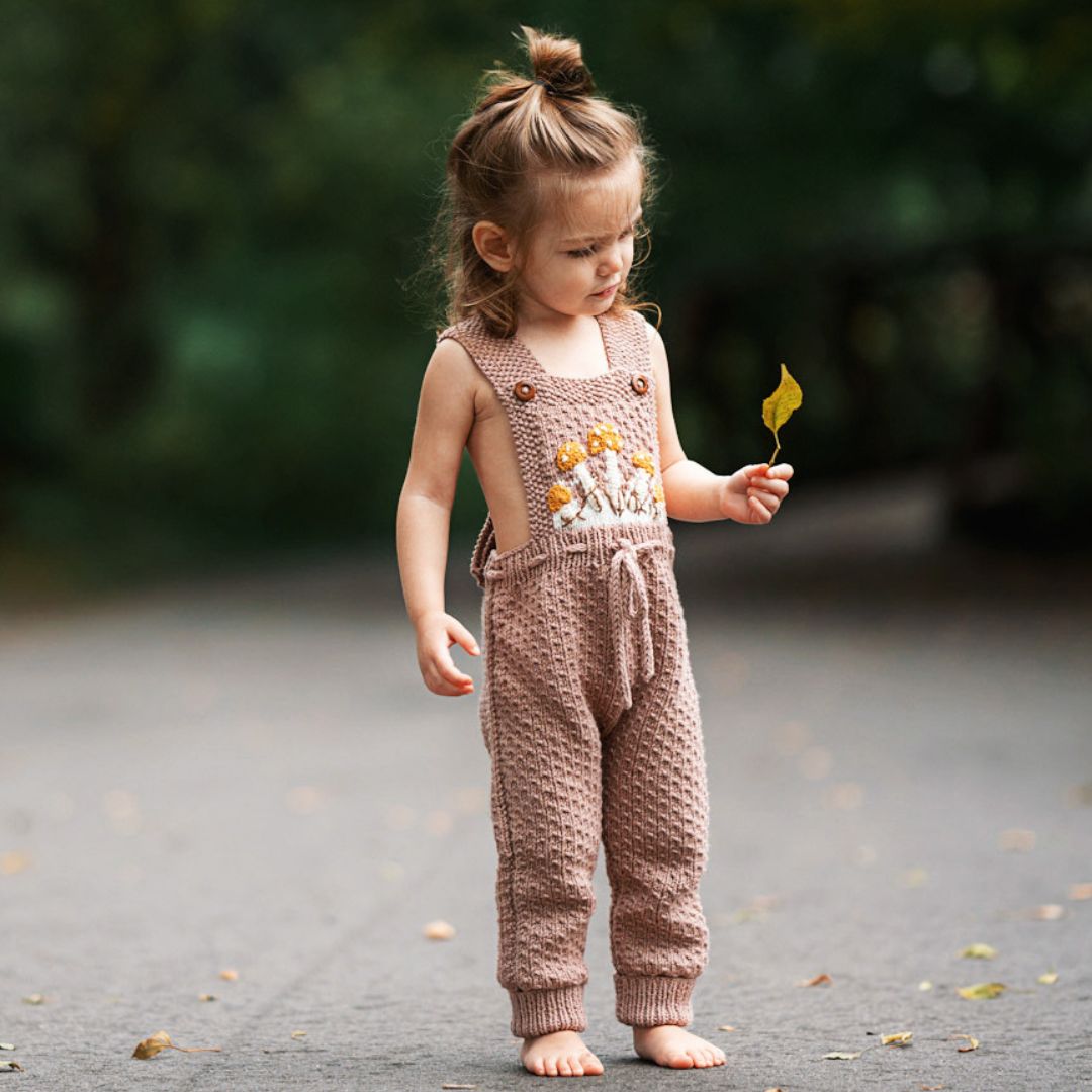 Toddler wearing hand-knit mushroom romper in blush brown cotton bamboo with adjustable criss-cross straps.