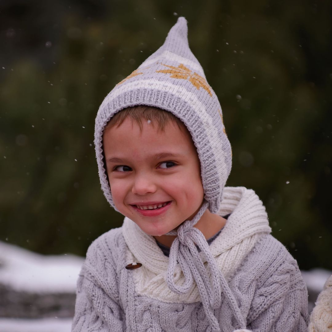 Child wearing unisex Nordic pixie hat in light gray hand-knit cotton bamboo with snowflake motif.