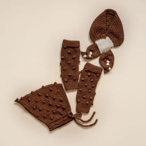 Brown popcorn knit pixie hat, scarf with cream accents, and matching leg warmers laid flat.

